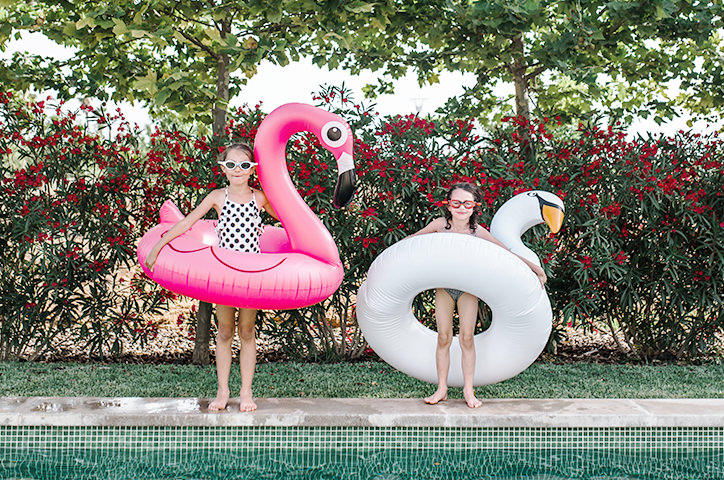 Two sisters in swimsuits and goggles with pool toys by the edge of the pool