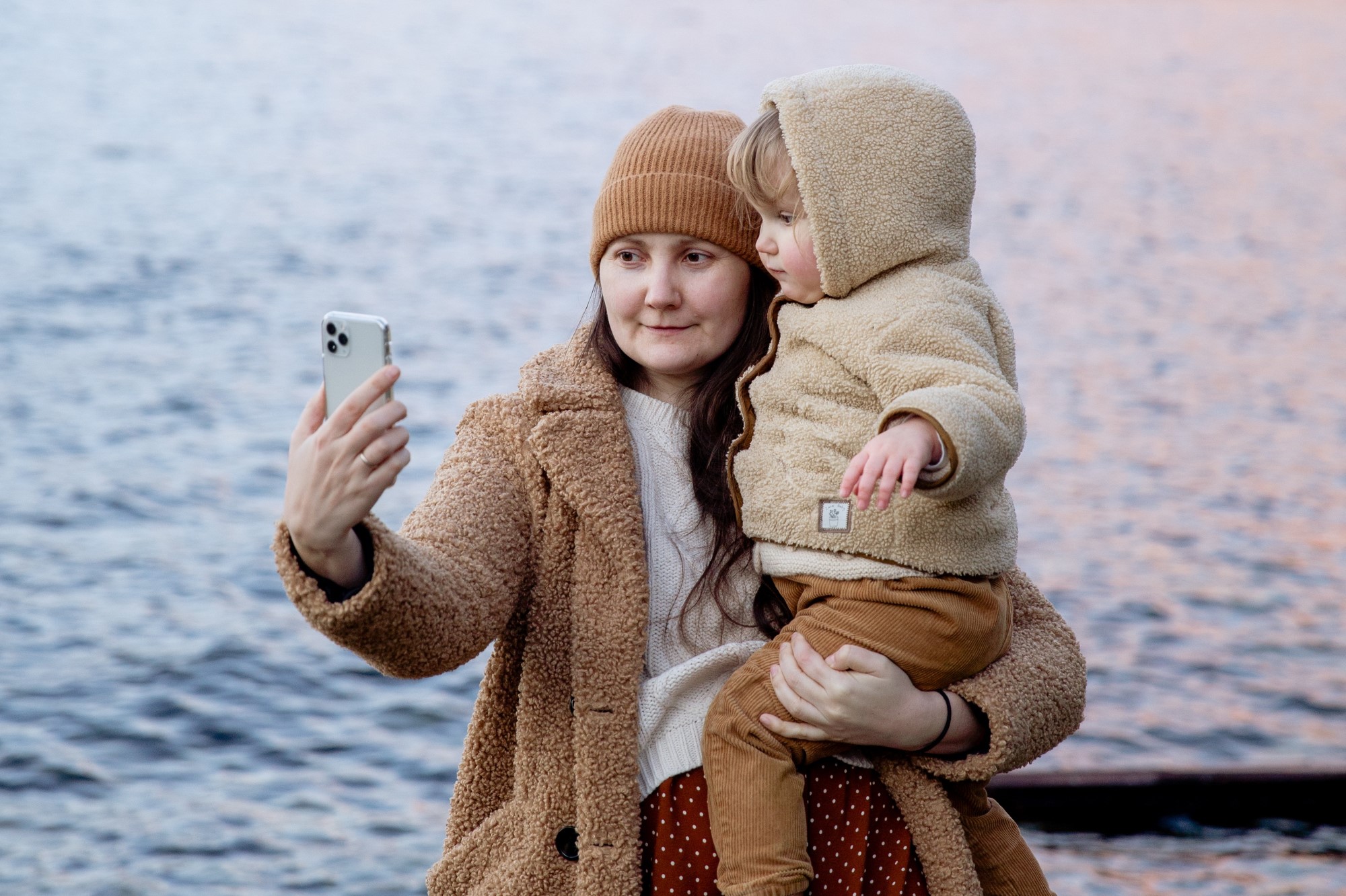 Woman wearing winter clothes holds baby for a phone selfie in front of calm water