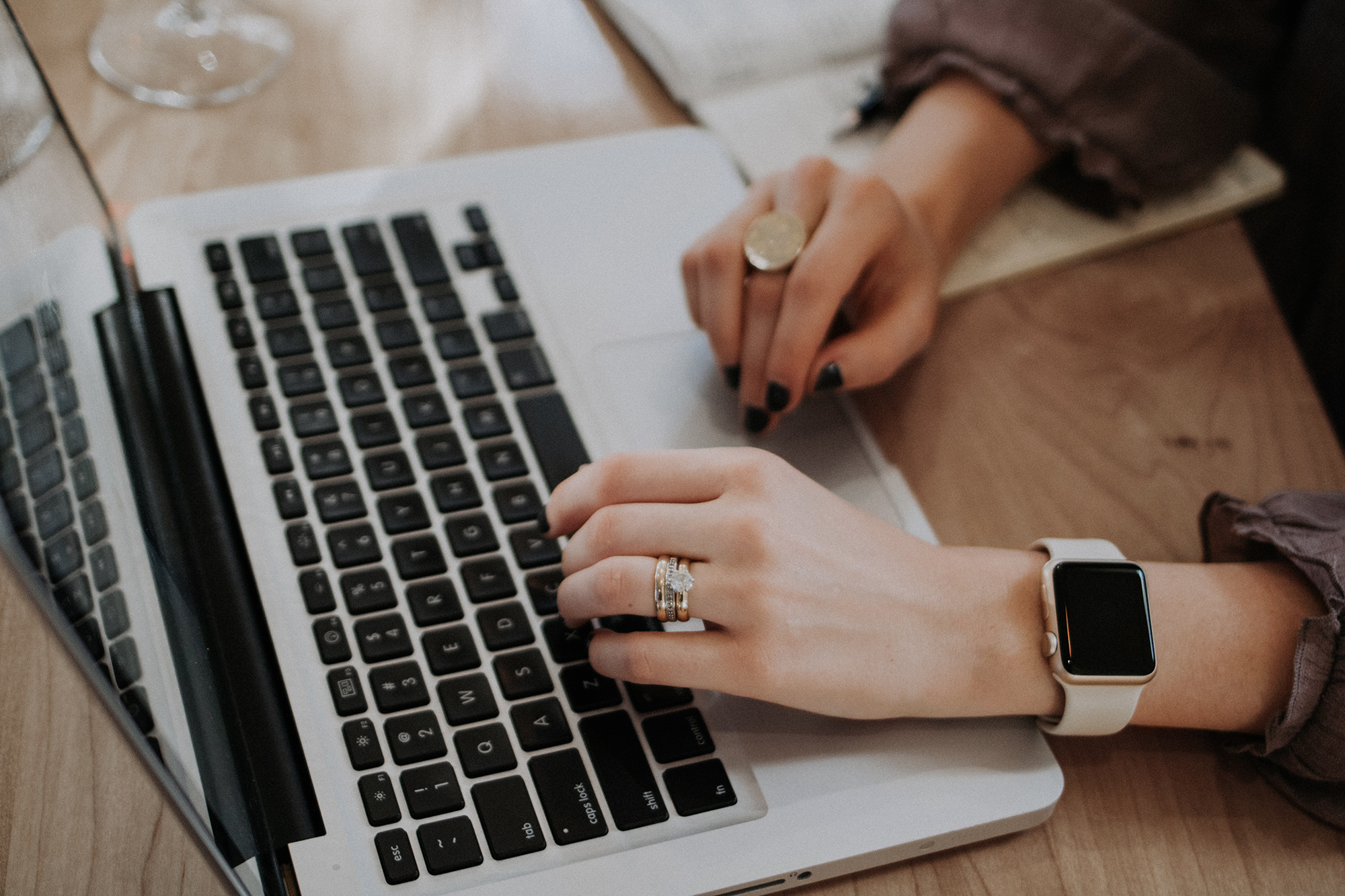 Womens hands on Apple Mac laptop sitting on a wooden table top, wearing rings and Apple Watch