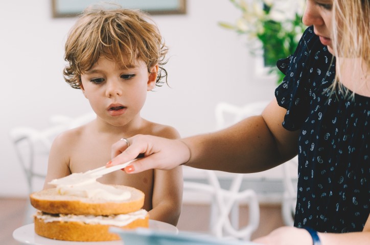 Mother spreads white icing on plain vanilla cake while young son watches