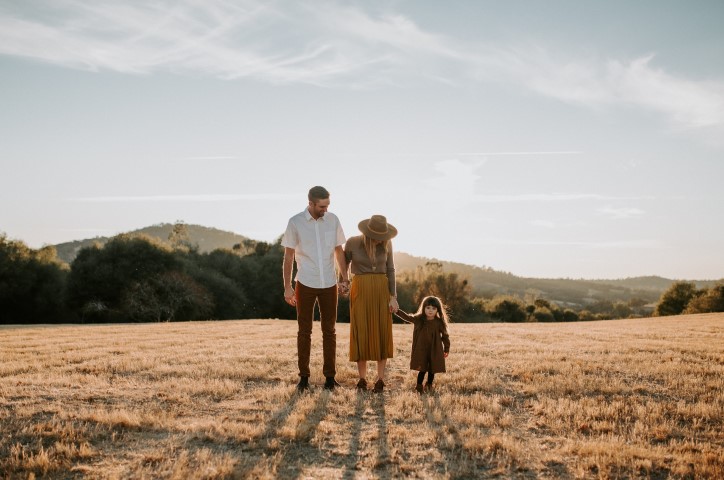 Family of three stand together holding hands in dried grass field in fall